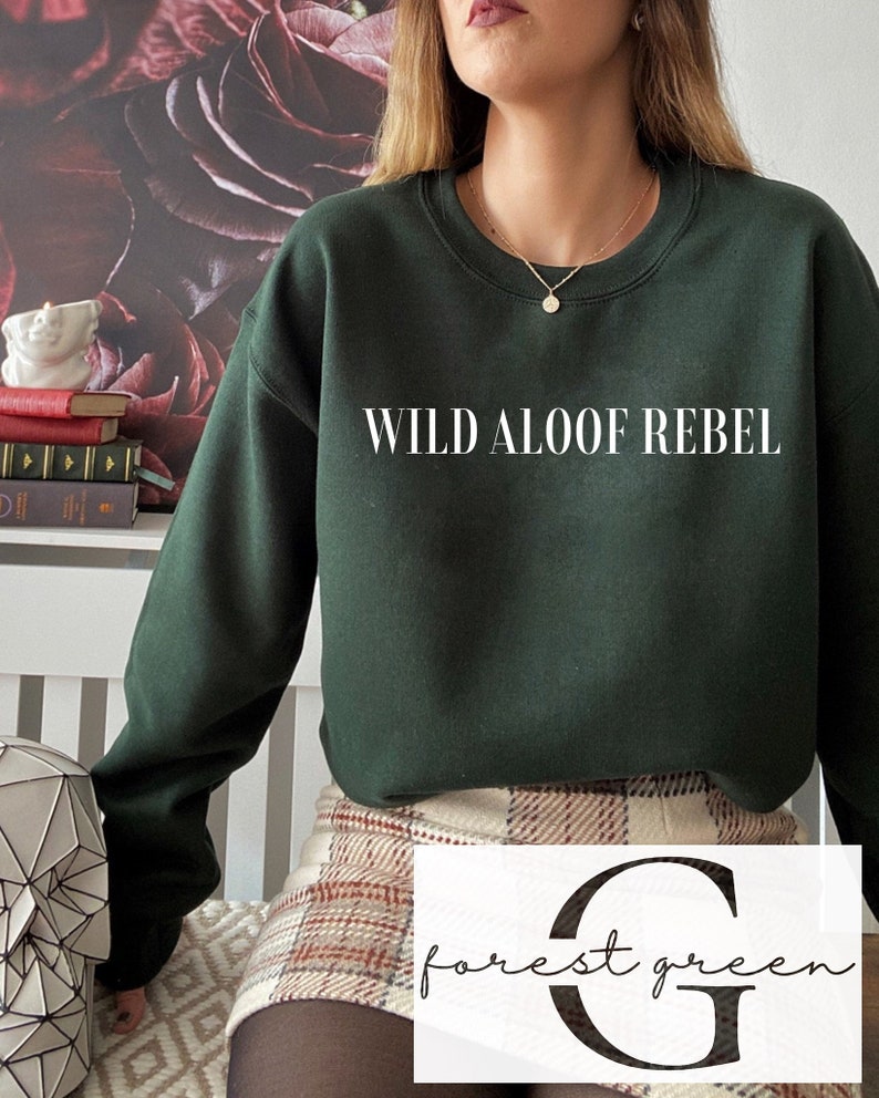 May include: A forest green sweatshirt with the text "WILD ALOOF REBEL" printed on the front. The sweatshirt is worn by a person wearing a plaid skirt.