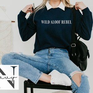 May include: A navy blue sweatshirt with the text "WILD ALOOF REBEL" printed on the front. The sweatshirt is being worn by a person sitting on a chair. The person is wearing blue jeans and white sneakers.