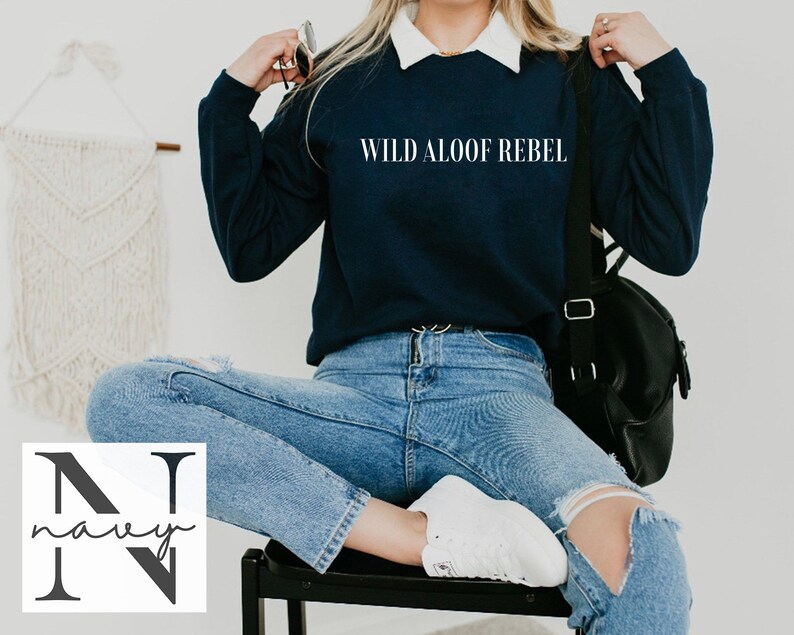 May include: A navy blue sweatshirt with the text "WILD ALOOF REBEL" printed on the front. The sweatshirt is being worn by a person sitting on a chair. The person is wearing blue jeans and white sneakers.