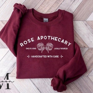 K&ouml;nnte beinhalten: Ein maroonfarbener Pullover mit dem Text "ROSE APOTHECARY" in Wei&szlig;, mit zwei stilisierten Rosen und den S&auml;tzen "ONE OF A KIND", "LOCALLY SOURCED" und "HANDCRAFTED WITH CARE".