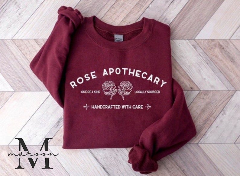 K&ouml;nnte beinhalten: Ein maroonfarbener Pullover mit dem Text "ROSE APOTHECARY" in Wei&szlig;, mit zwei stilisierten Rosen und den S&auml;tzen "ONE OF A KIND", "LOCALLY SOURCED" und "HANDCRAFTED WITH CARE".