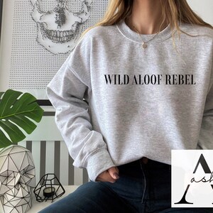 May include: A gray sweatshirt with the text "WILD ALOOF REBEL" printed on the front. The sweatshirt is being worn by a person with dark hair and dark jeans.