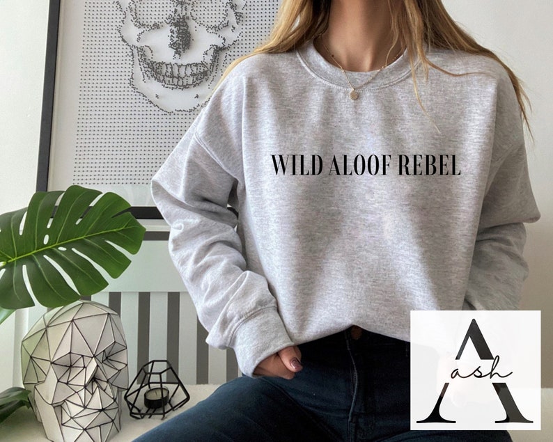 May include: A gray sweatshirt with the text "WILD ALOOF REBEL" printed on the front. The sweatshirt is being worn by a person with dark hair and dark jeans.