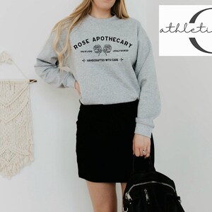 K&ouml;nnte beinhalten: Ein graues Sweatshirt mit dem Text "ROSE APOTHECARY" und einem floralen Design. Das Sweatshirt wird von einer Person getragen, die einen schwarzen Rock tr&auml;gt und einen schwarzen Rucksack h&auml;lt.