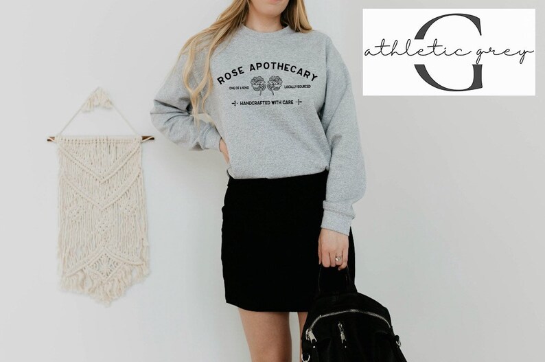 K&ouml;nnte beinhalten: Ein graues Sweatshirt mit dem Text "ROSE APOTHECARY" und einem floralen Design. Das Sweatshirt wird von einer Person getragen, die einen schwarzen Rock tr&auml;gt und einen schwarzen Rucksack h&auml;lt.