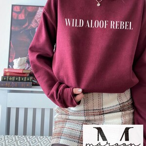 May include: A maroon sweatshirt with the text "WILD ALOOF REBEL" printed in white on the front. The sweatshirt is worn by a person wearing a plaid skirt.