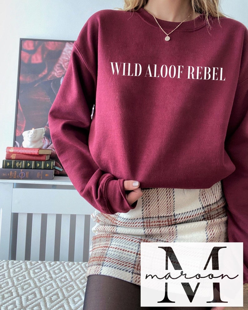 May include: A maroon sweatshirt with the text "WILD ALOOF REBEL" printed in white on the front. The sweatshirt is worn by a person wearing a plaid skirt.
