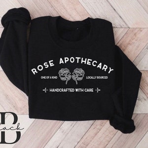 K&ouml;nnte beinhalten: Schwarzer Pullover mit wei&szlig;em Text, der "Rose Apothecary" lautet, mit zwei Rosen und den Worten "One of a Kind" und "Locally Sourced". Der Text "Handcrafted with Care" ist ebenfalls enthalten.