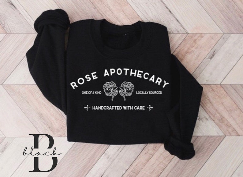 K&ouml;nnte beinhalten: Schwarzer Pullover mit wei&szlig;em Text, der "Rose Apothecary" lautet, mit zwei Rosen und den Worten "One of a Kind" und "Locally Sourced". Der Text "Handcrafted with Care" ist ebenfalls enthalten.