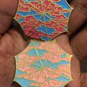 May include: Two gold-toned enamel pins shaped like umbrellas with a blue and pink cloud design. The clouds are filled with glitter.