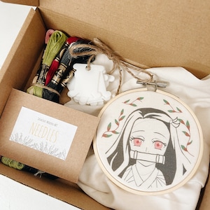 May include: An open cardboard box containing an embroidery kit. The kit includes embroidery floss, a wooden hoop with a character design, a needle packet, and a white tag. The character design features a girl with pink eyes and a bamboo muzzle.