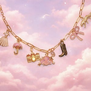 May include: Gold-toned charm necklace featuring various miniature charms, including a dress, umbrella, camera, cloud, cowboy boot, bow, and a winged horse. The necklace is set against a soft pink and white cloudy background.