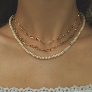 Puede incluir: Tres collares de oro superpuestos en el cuello de una persona. El collar superior tiene peque&ntilde;as piedras de colores. El collar del medio tiene piedras ovaladas m&aacute;s grandes. El collar inferior est&aacute; hecho de peque&ntilde;as cuentas blancas.