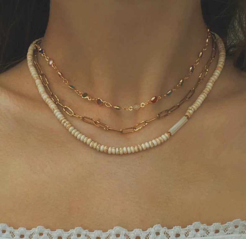 Puede incluir: Tres collares de oro superpuestos en el cuello de una persona. El collar superior tiene peque&ntilde;as piedras de colores. El collar del medio tiene piedras ovaladas m&aacute;s grandes. El collar inferior est&aacute; hecho de peque&ntilde;as cuentas blancas.