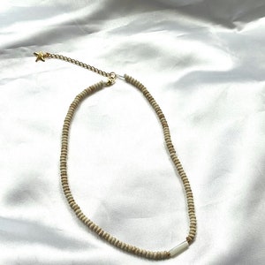 Puede incluir: Un collar de cuentas blancas con un dije de estrella dorada y un acento de concha blanca. El collar est&aacute; sobre un fondo de tela blanca.