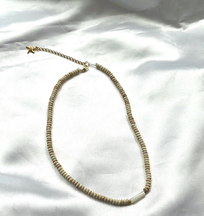 Puede incluir: Un collar de cuentas blancas con un dije de estrella dorada y un acento de concha blanca. El collar est&aacute; sobre un fondo de tela blanca.