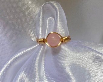 May include: A delicate gold wire ring with a pink gemstone. The ring is on a white satin background.