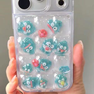 Puede incluir: Funda transparente para teléfono con una cuadrícula de personajes de dibujos animados azules y detalles rojos. La funda tiene un borde transparente y un recorte para la cámara. Los personajes de dibujos animados tienen diversas expresiones y diseños.