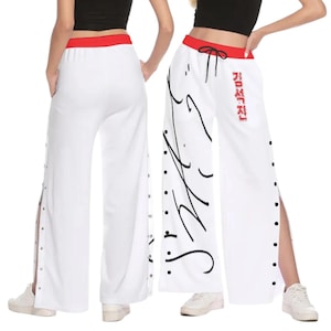 Puede incluir: Pantalones blancos de pernera ancha con cintura roja y aberturas laterales. Los pantalones presentan escritura negra y texto coreano en una pernera y cierres de presión plateados a lo largo de las costuras laterales.