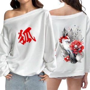 Può includere: Felpa bianca con spalle scoperte. La parte anteriore presenta un carattere cinese rosso. Il retro mostra un disegno ad acquerello di una volpe con pelliccia rossa e bianca, circondata da fiori rossi e foglie grigie. Design artistico e casual.