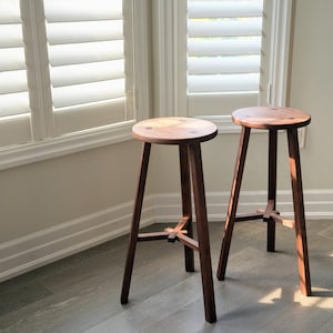 May include: Two wooden stools with a simple, rustic design. The stools have a round seat and four legs that form an X shape at the base.