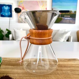 May include: A glass coffee maker with a wooden collar and a leather strap. The coffee maker has a glass carafe and a stainless steel filter. The carafe has markings for 200, 300, and 400 millilitres.