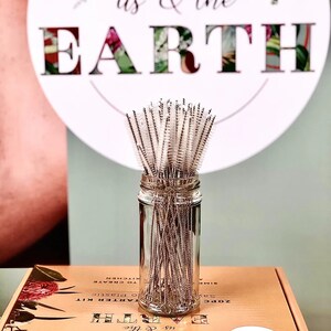 May include: A glass jar filled with a bundle of stainless steel cleaning brushes with white bristles. The jar is sitting on a box with the text "us & the EARTH" printed on it.
