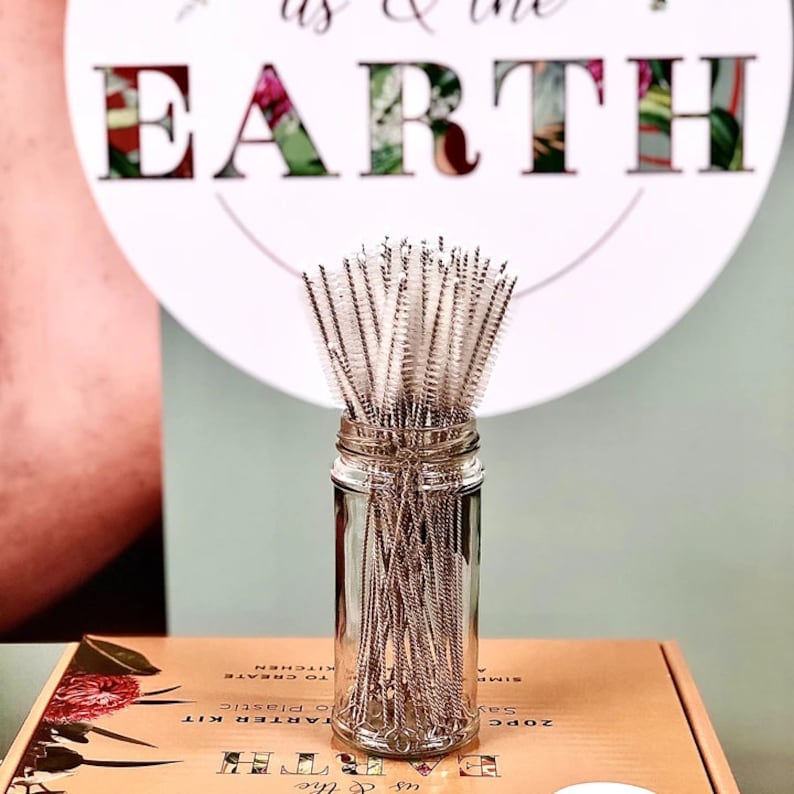 May include: A glass jar filled with a bundle of stainless steel cleaning brushes with white bristles. The jar is sitting on a box with the text "us & the EARTH" printed on it.