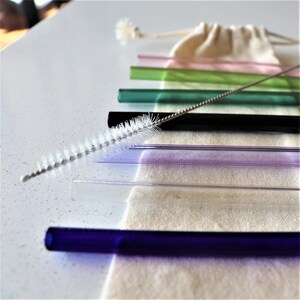 May include: A set of colourful glass drinking straws with a cleaning brush. The straws are in a variety of colours, including pink, green, black, purple, clear, and blue. The straws are arranged in a row on a white surface.
