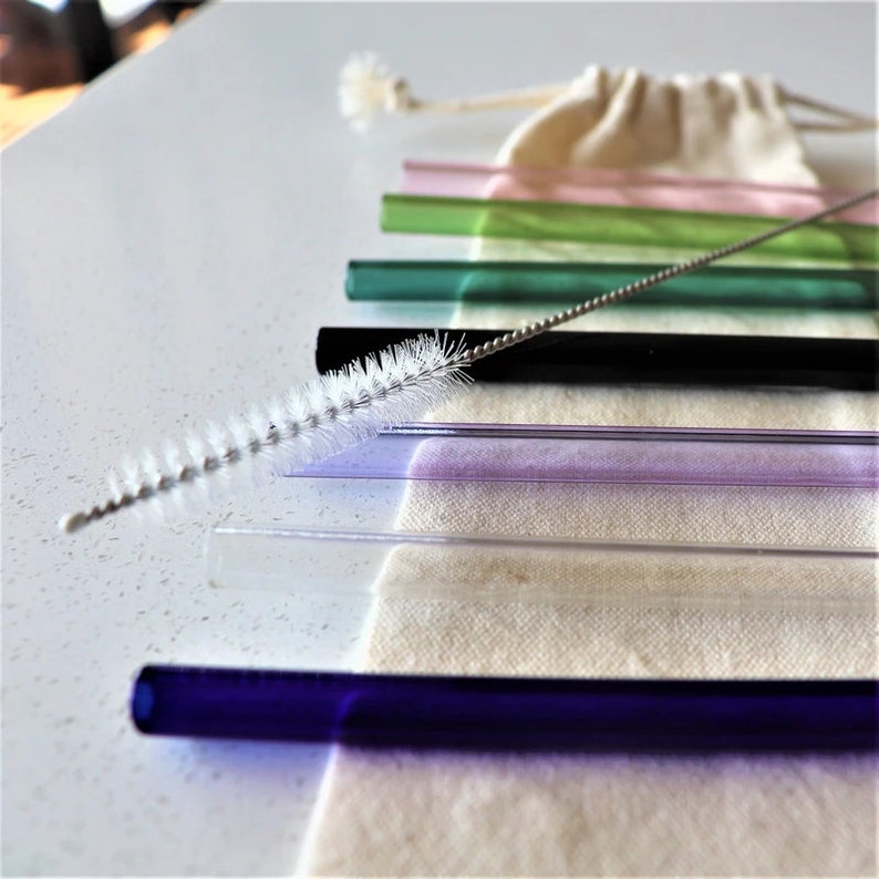 May include: A set of colourful glass drinking straws with a cleaning brush. The straws are in a variety of colours, including pink, green, black, purple, clear, and blue. The straws are arranged in a row on a white surface.
