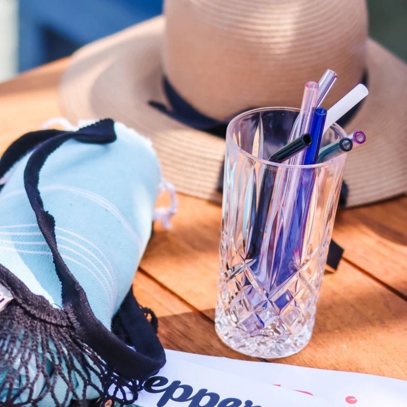 May include: A glass of water with a selection of colourful reusable drinking straws. The straws are made of glass and come in a range of colours, including blue, purple, and pink. The glass is clear and has a diamond pattern.