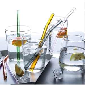 May include: Four clear glass tumblers filled with water and lemon slices. Each tumbler has a different coloured glass straw. The straws are green, yellow, pink and purple.