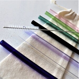 May include: A set of colourful glass drinking straws with a cleaning brush. The straws are in a variety of colours, including blue, purple, green, and pink. The straws are arranged in a row on a white surface.