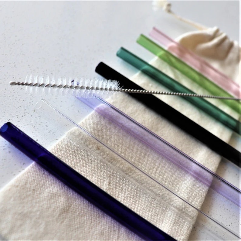 May include: A set of colourful glass drinking straws with a cleaning brush. The straws are in a variety of colours, including blue, purple, green, and pink. The straws are arranged in a row on a white surface.