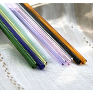 May include: A set of seven glass drinking straws in various colours, including green, blue, yellow, white, lavender, black, and orange. The straws are arranged in a row on a white fabric surface.