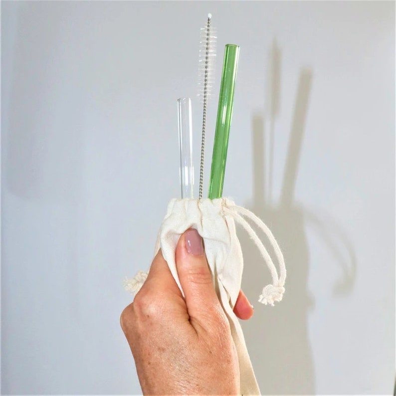 May include: A set of two glass drinking straws, one clear and one green, with a cleaning brush, all stored in a white cotton drawstring bag.