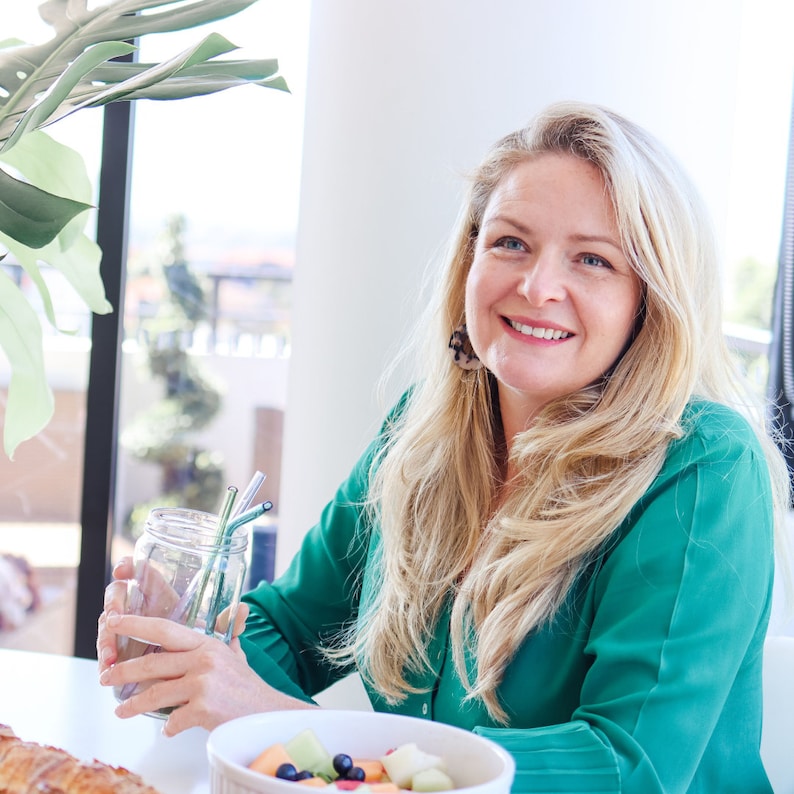 May include: A woman with long blonde hair is sitting at a table with a glass jar and a bowl of fruit. She is holding a glass jar with a green straw in it. The jar is filled with a clear liquid. The bowl of fruit is filled with fresh fruit, including melon, berries, and kiwi.