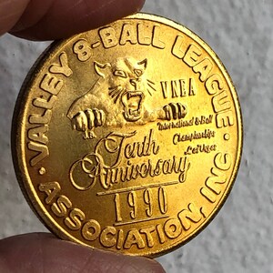 May include: A gold-coloured coin with the text "Valley 8-Ball League Association, Inc." and "Tenth Anniversary 1990" surrounding a cartoon image of a snarling cat. The text "VNEA International 8-Ball Championships Las Vegas" is also included.