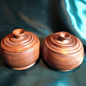 May include: Two wooden lidded containers with a dark brown finish. The containers have a rounded shape and are made of wood with a smooth, polished surface.