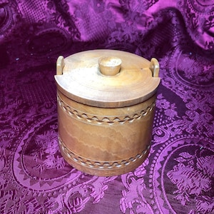 May include: A round wooden box with a carved design and a lid. The box is made of light brown wood and has a natural finish. The lid has a small knob on top.