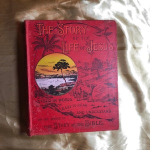 May include: A red book titled "The Story of the Life of Jesus" with a yellow sunset scene on the cover. The book is written in words easy to read and understand by the author of "The Story of the Bible."