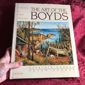 May include: A book titled "The Art of the Boyds: Generations of Artistic Achievement" by Patricia Dobrez and Peter Herbst. The cover features a painting of a coastal scene with a boat under construction and a group of people working on it.