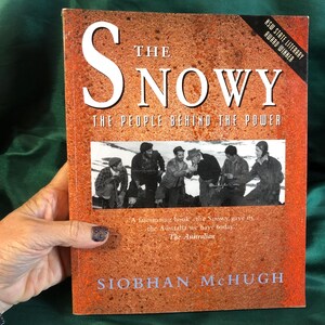 May include: A book titled "The Snowy: The People Behind the Power" by Siobhan McHugh. The cover is orange-brown with white text and a black and white photograph of men. It has a sticker that says "NSW State Literary Award Winner".