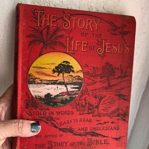 May include: Red book cover with the title "The Story of the Life of Jesus" in large, ornate lettering. The cover features illustrations of a palm tree, a sunset over a lake, and a group of people with camels. The text below the title reads "Told in Words Easy to Read and Understand. By the Author of The Story of the Bible."