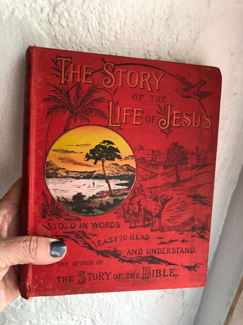 May include: Red book cover with the title "The Story of the Life of Jesus" in large, ornate lettering. The cover features illustrations of a palm tree, a sunset over a lake, and a group of people with camels. The text below the title reads "Told in Words Easy to Read and Understand. By the Author of The Story of the Bible."