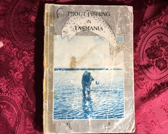 Pode incluir: Uma capa de livro vintage com fundo marrom e uma imagem de uma truta saltando para fora da água. O título do livro é "Trout Fishing in Tasmania" de Greg French.