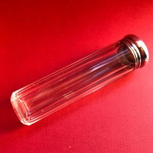 May include: A clear, rectangular glass vial with vertical ridges and a silver-coloured metal cap. The vial is lying on a red surface. The cap has a decorative, ridged design.