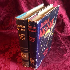 May include: Two vintage hardcover books by Alistair MacLean. The book spines are dark blue and brown with gold lettering. The titles are "When Eight Bells Toll" and "Where Eagles Dare". The book covers show signs of wear.