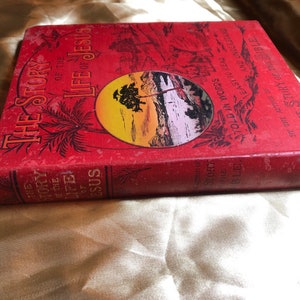 May include: A red hardcover book titled "The Story of the Life of Jesus" with gold lettering. The book cover features a sunset scene with a palm tree and a body of water. The text on the cover reads "Told in Words Easy to Read and Understand" and "By the Author of The Story of the Bible."