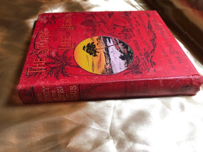 May include: A red hardcover book titled "The Story of the Life of Jesus" with gold lettering. The book cover features a sunset scene with a palm tree and a body of water. The text on the cover reads "Told in Words Easy to Read and Understand" and "By the Author of The Story of the Bible."